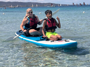 Woman and boy smiling while paddling on a board in the sea, both wearing life jackets and representing children with autism.